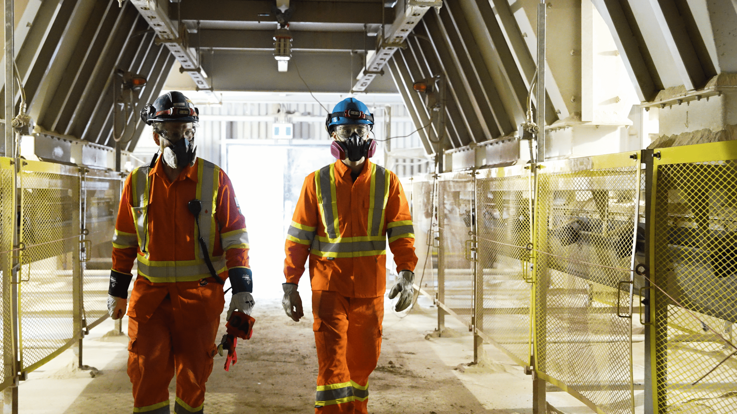 Cement and Concrete Solutions - two workers in orange coveralls and respirators walking through a plant walkway